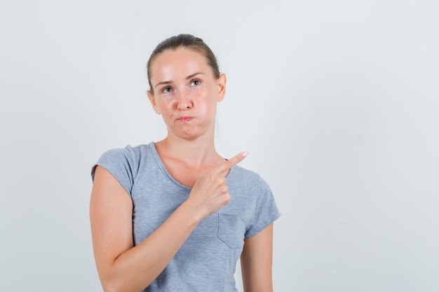 Jeune femme en t-shirt gris pointant vers le côté et regardant mécontent, vue de face.