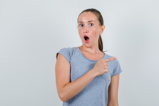 Jeune femme en t-shirt gris pointant vers le côté et à la surprise, vue de face.