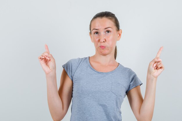 Jeune femme en t-shirt gris pointant vers le haut et à la perplexité, vue de face.