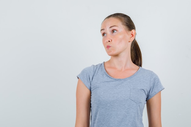 Jeune femme en t-shirt gris à la recherche de loin et à la vue de face, hésitante.