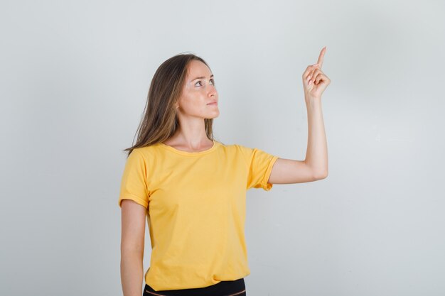 Jeune femme en t-shirt jaune, pantalon pointant le doigt vers le haut et en détournant les yeux