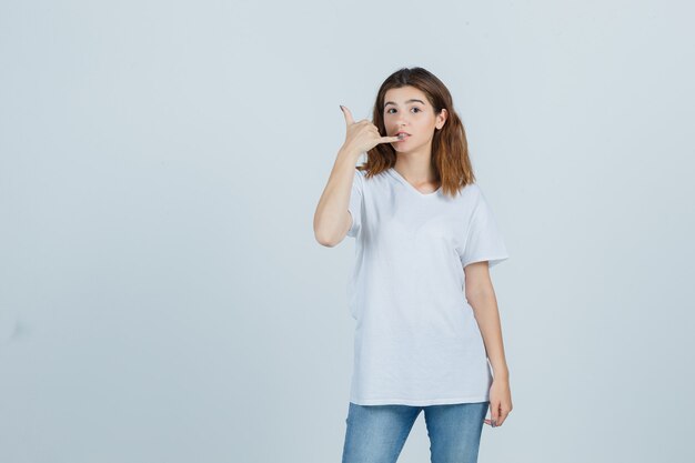 Jeune femme en t-shirt, jeans montrant le geste du téléphone et l'air confiant, vue de face.