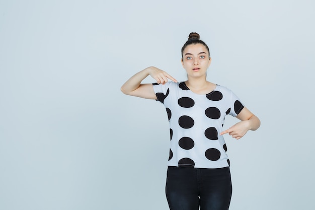 Jeune femme en t-shirt, jeans pointant vers l'extérieur et regardant perplexe, vue de face.