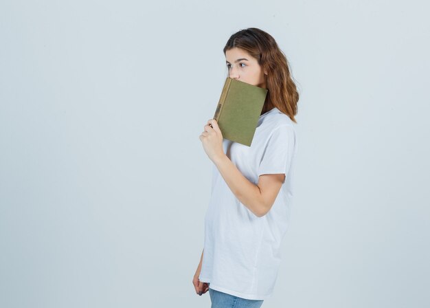 Jeune femme en t-shirt, jeans s'appuyant sur le livre sur la bouche et à la vue réfléchie, de face.
