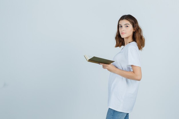 Jeune femme en t-shirt, jeans tenant un livre tout en regardant par-dessus l'épaule et à la vue de face, confiant.