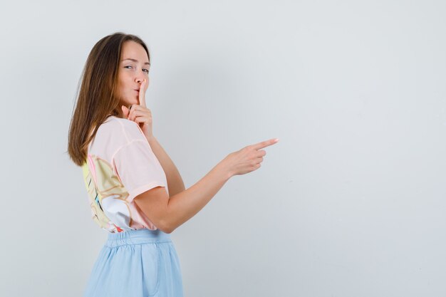 Jeune femme en t-shirt, jupe pointant vers quelque chose avec un geste de silence.