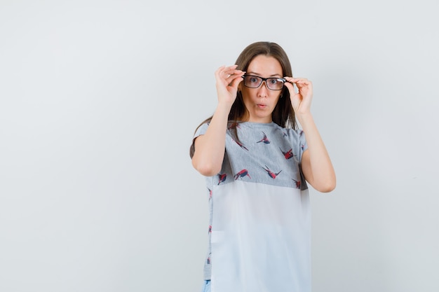 Jeune femme en t-shirt, jupe regardant attentivement à travers des lunettes et regardant étonné, vue de face.