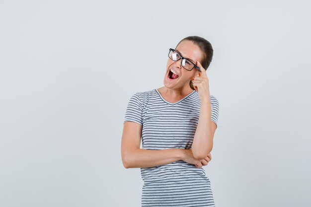 Jeune femme en t-shirt, lunettes bâillant tout en regardant ailleurs et à la pensive, vue de face.