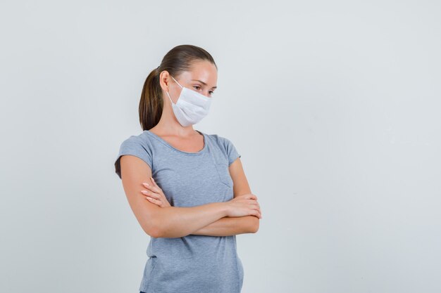 Jeune femme en t-shirt, masque debout avec les bras croisés et regardant pensif, vue de face.