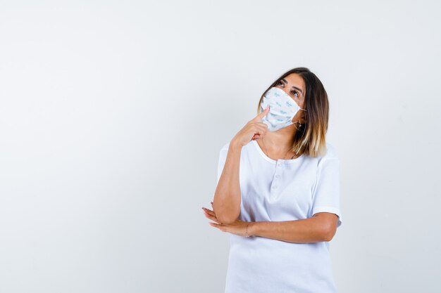 Jeune femme en t-shirt, masque debout dans la pose de pensée et à la vue réfléchie, de face.