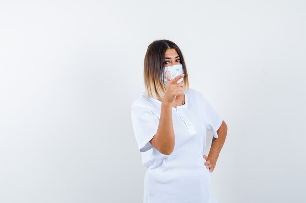 Jeune femme en t-shirt, masque pointant de côté tout en gardant la main sur la taille et à la vue de face confiante.