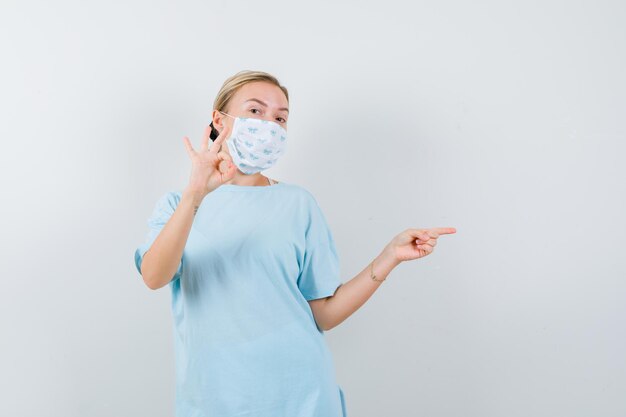 Jeune femme en t-shirt, masque pointant vers le côté droit tout en montrant le signe ok