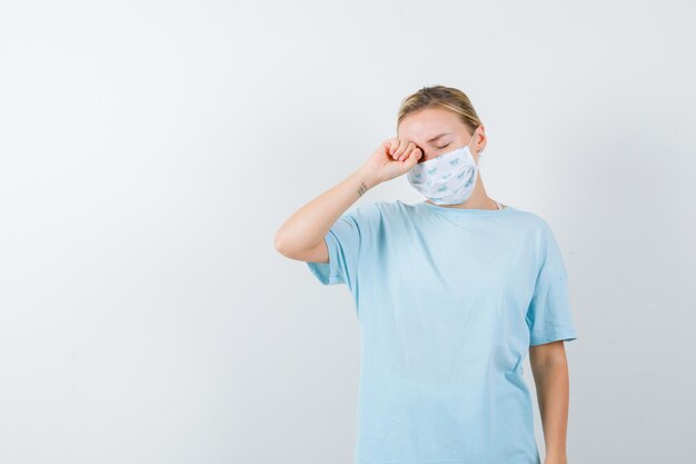 Jeune femme en t-shirt, masque se frottant les yeux avec le poing et ayant l'air endormi