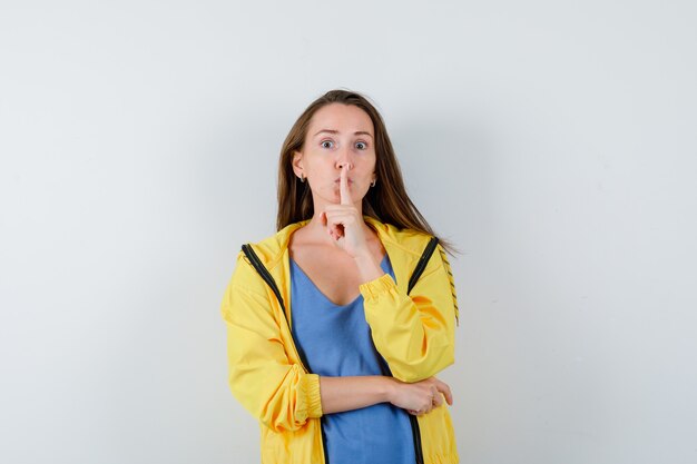 Jeune femme en t-shirt montrant un geste de silence et regardant attentivement, vue de face.