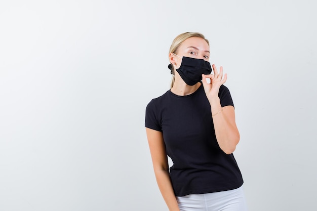 Jeune femme en t-shirt, pantalon, masque médical montrant un geste de fermeture éclair et l'air sérieux