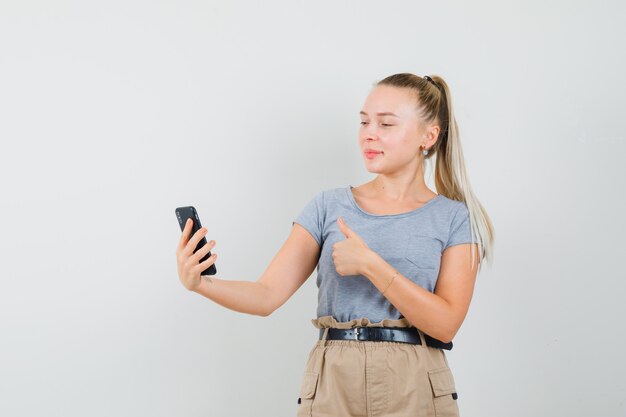 Jeune femme en t-shirt, pantalon montrant le pouce vers le haut sur le chat vidéo et à la jolly, vue de face.