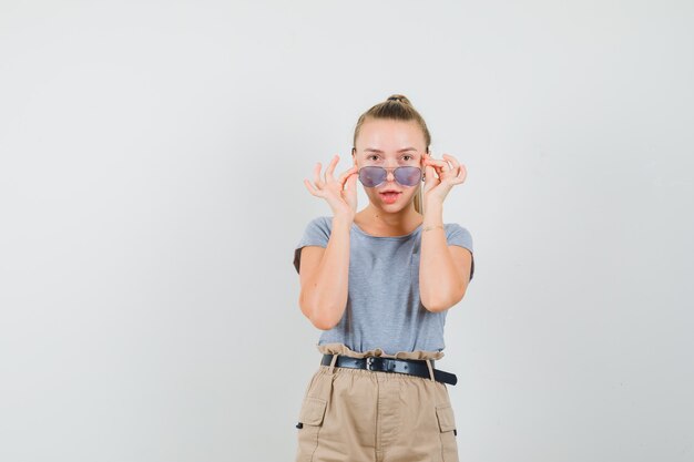 Jeune femme en t-shirt, pantalon regardant attentivement par-dessus les lunettes et à la vue de face, focalisée.
