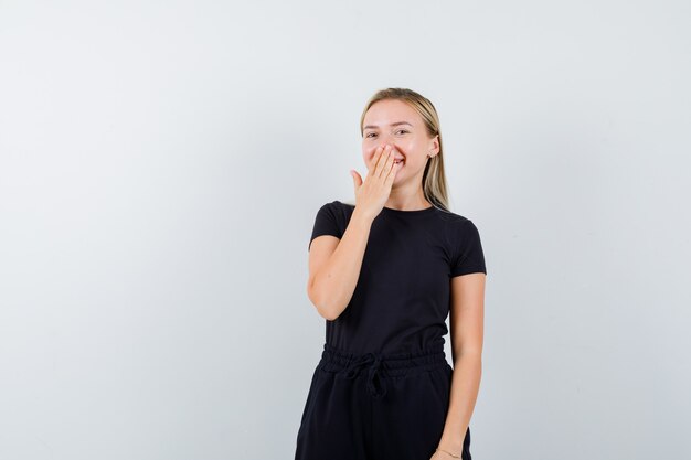 Jeune femme en t-shirt, pantalon tenant la main sur la bouche et l'air heureux, vue de face.