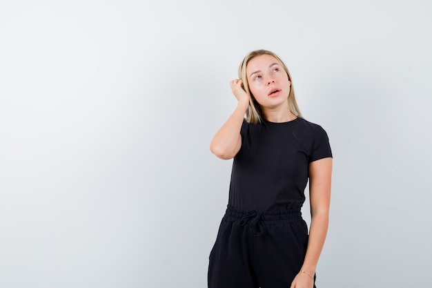 Jeune femme en t-shirt, pantalon tenant la main sur la tête et à la vue réfléchie, de face.