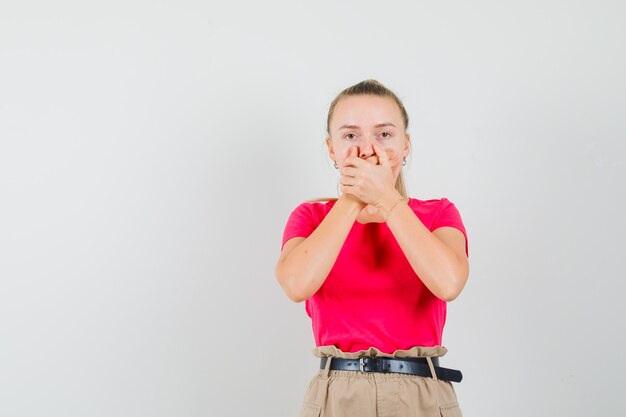 Jeune femme en t-shirt, pantalon tenant les mains sur la bouche et à la surprise, vue de face.