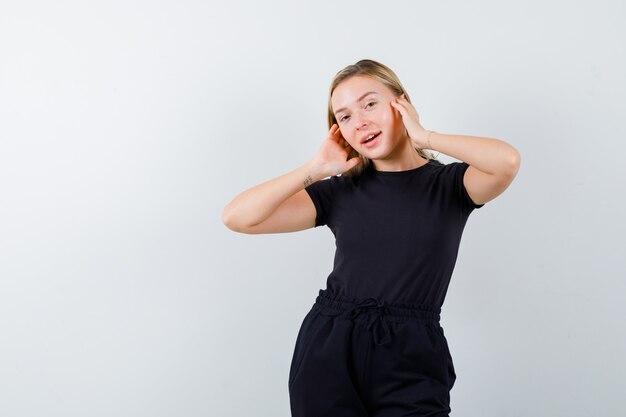 Jeune femme en t-shirt, pantalon tenant les mains sur les oreilles et à la joyeuse vue de face.