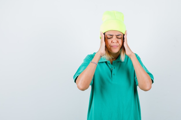 Jeune femme en t-shirt polo, bonnet avec les mains près du visage, fermant les yeux et regardant concentré, vue de face.