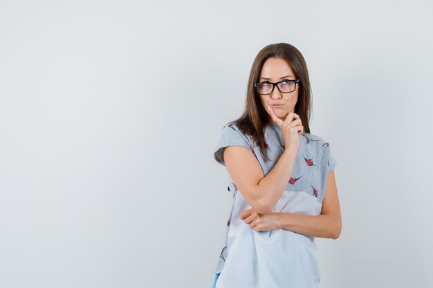 Jeune femme en t-shirt à la recherche avec le doigt sur le menton et à la pensive, vue de face.