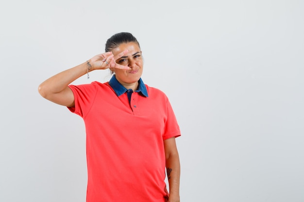 Jeune femme en t-shirt rouge montrant le signe v sur les yeux et l'air heureux, vue de face.