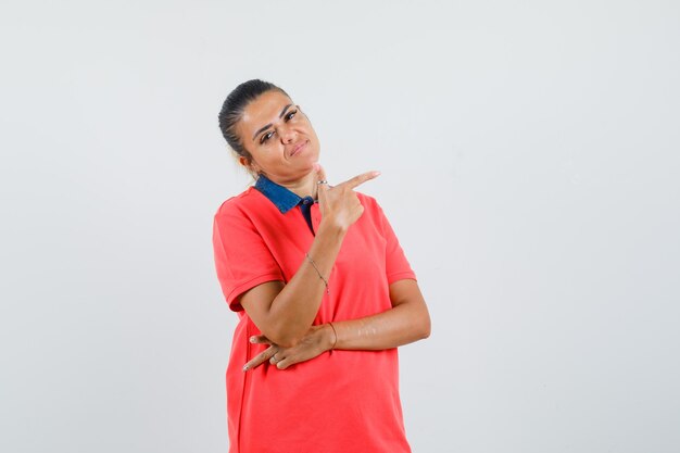 Jeune femme en t-shirt rouge pointant vers la droite avec l'index et à la jolie vue de face.