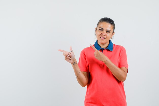 Jeune femme en t-shirt rouge pointant vers la gauche avec l'index et l'air mécontent, vue de face.