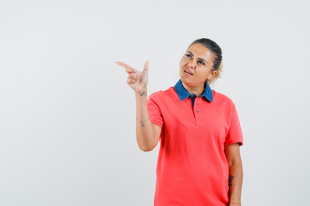 Jeune femme en t-shirt rouge pointant vers la gauche et à la vue ennuyée, de face.