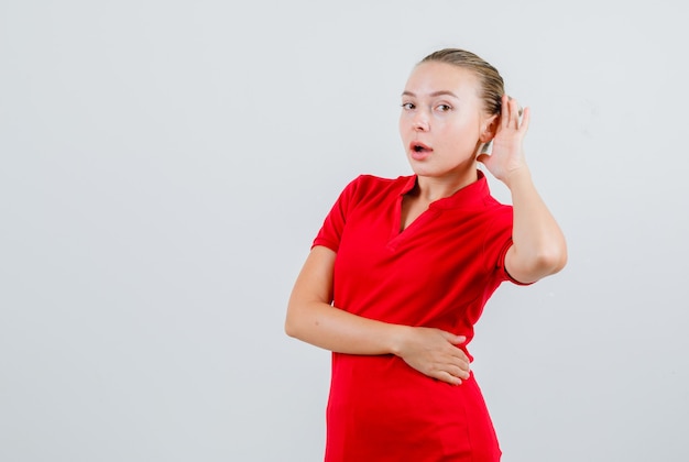 Jeune femme en t-shirt rouge tenant la main derrière l'oreille et à la curiosité