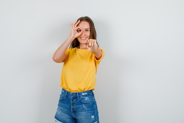 Jeune femme en t-shirt, short pointant la caméra avec signe ok sur les yeux et à la bonne humeur