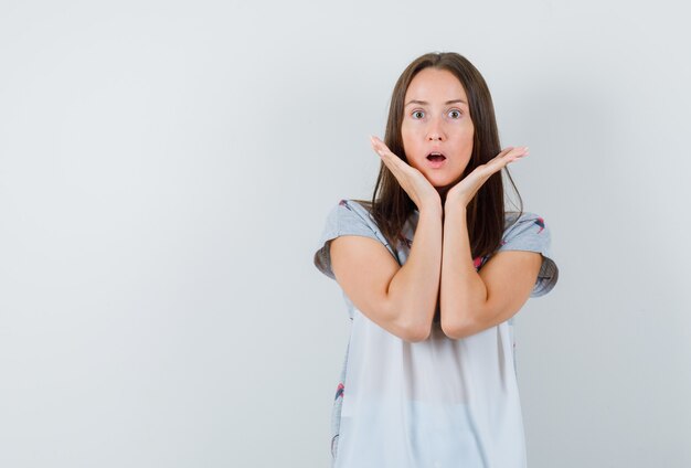 Jeune femme en t-shirt tenant les paumes surélevées près du menton et à la surprise, vue de face.