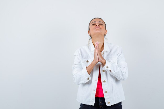 Jeune femme en t-shirt, veste blanche avec les mains en geste de prière et l'air plein d'espoir, vue de face.