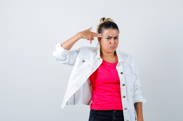 Jeune femme en t-shirt, veste blanche montrant le geste du pistolet et semblant s'ennuyer, vue de face.