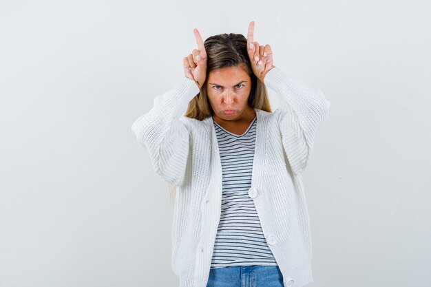 Jeune femme en t-shirt, veste en gardant les doigts sur la tête comme des cornes de taureau et à la drôle, vue de face.