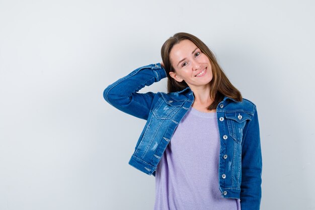 Jeune femme en t-shirt, veste en jean gardant la main derrière la tête et l'air joyeux, vue de face.