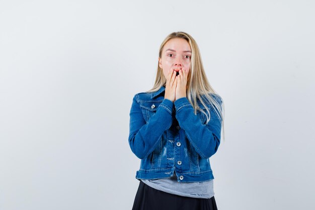 Jeune femme en t-shirt, veste en jean, jupe couvrant la bouche avec les mains et l'air étonné