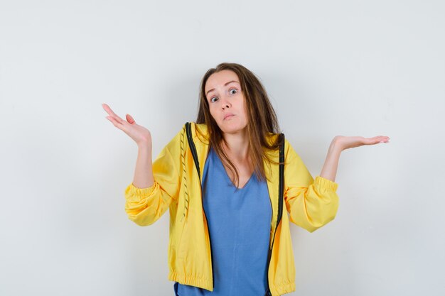 Jeune femme en t-shirt, veste montrant un geste impuissant en haussant les épaules et l'air confus, vue de face.