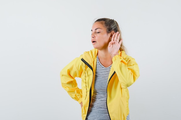 Jeune femme en t-shirt, veste tenant la main près de l'oreille et regardant curieux, vue de face.