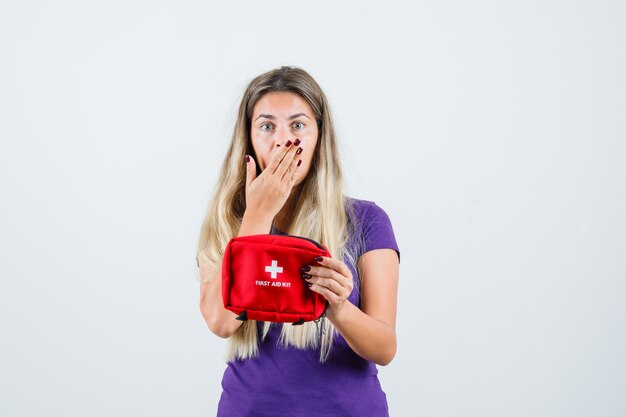 Jeune femme en t-shirt violet tenant une trousse de premiers soins et l'air inquiet, vue de face.