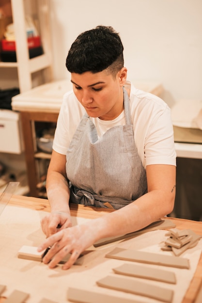 Jeune femme en tablier coupant l&#39;argile avec des outils sur une table en bois