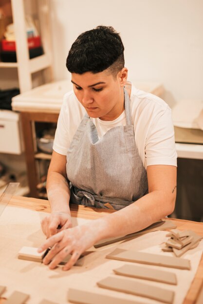 Jeune femme en tablier coupant l&#39;argile avec des outils sur une table en bois