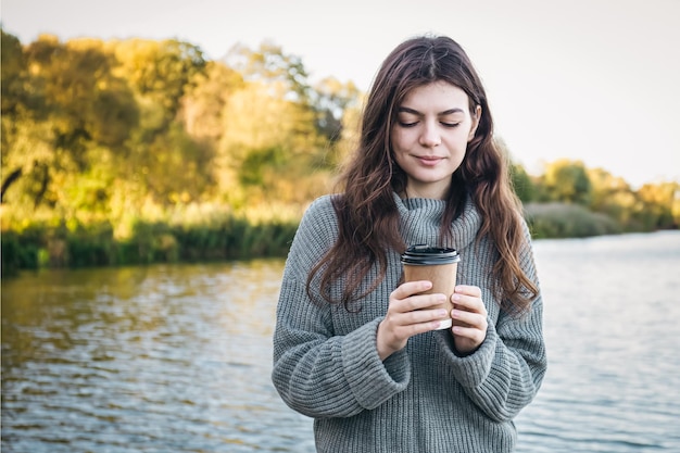 Photo gratuite une jeune femme avec une tasse de café sur un arrière-plan flou de la rivière