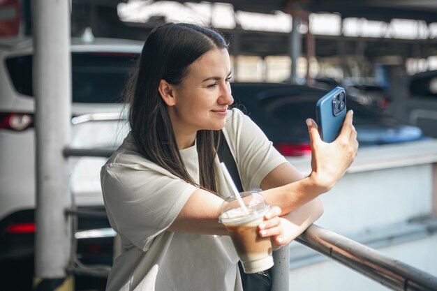 Une jeune femme avec une tasse de café dans le parking prend un selfie