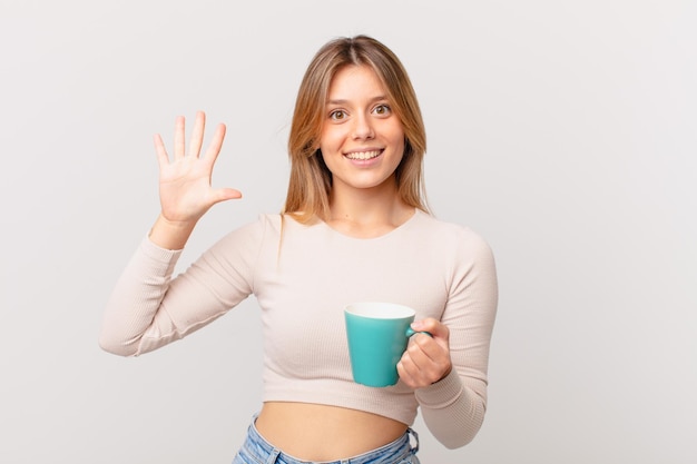 Jeune femme avec une tasse de café souriante et amicale, montrant le numéro cinq