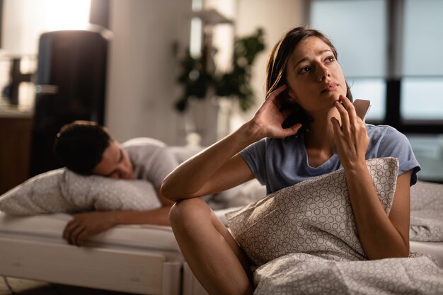 Jeune femme avec un téléphone intelligent contemplant et s'inquiétant de sa relation pendant que son petit ami dort sur le lit