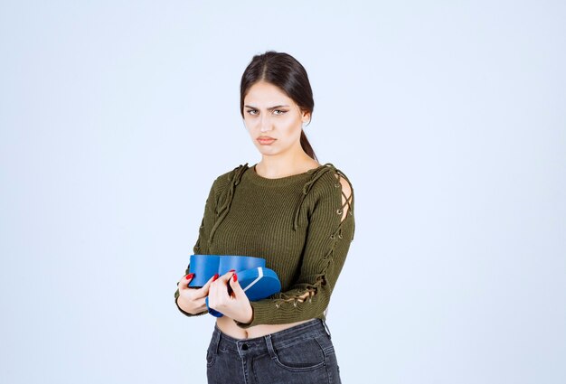 Jeune femme tenant une boîte cadeau bleue avec une expression de colère sur fond blanc.