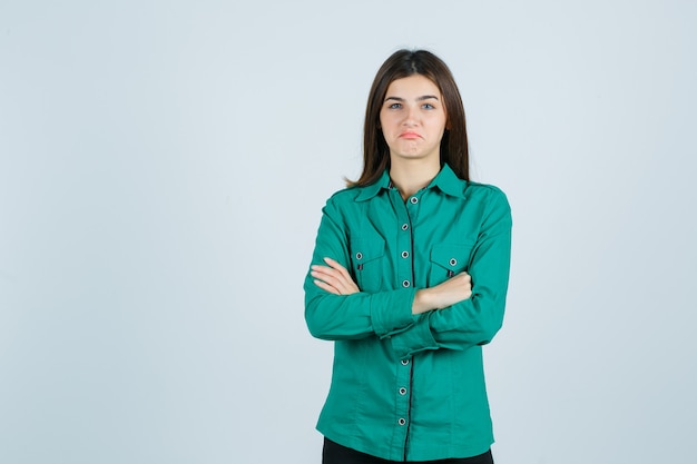 Jeune femme tenant les bras croisés tout en courbant les lèvres en chemise verte et à mécontent, vue de face.